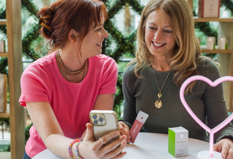 Two women smiling at each other and looking at a phone while holding Neora’s Slim + Skin Collagen Powder